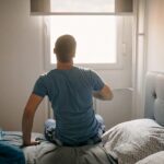 Man sitting on the edge of a bed, facing a bright window, reflecting on personal health decisions after a herpes outbreak.